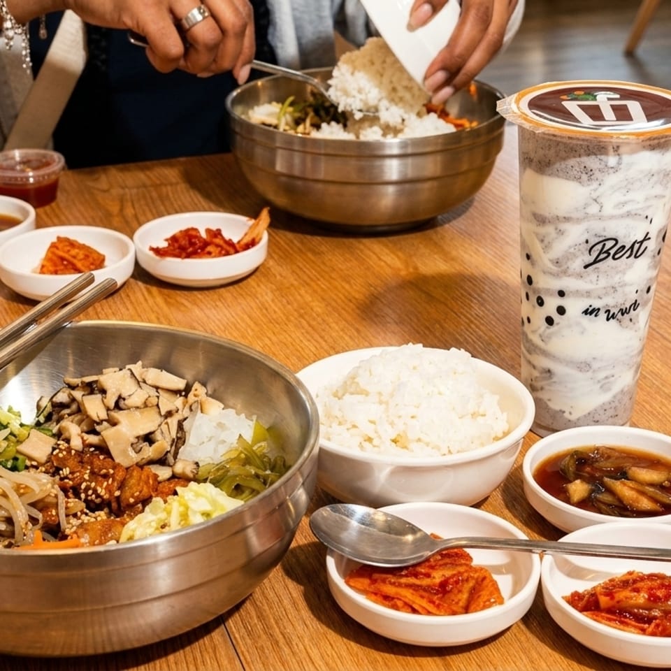 A person adding rice to a large bowl of bibimbap, surrounded by side dishes and a drink.